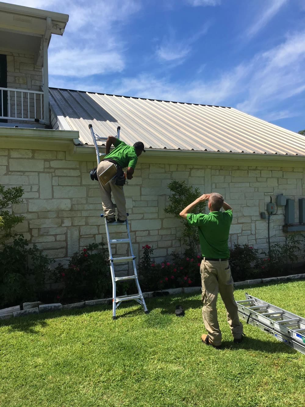 Two workers in green shirts beside a ladder on a stone house with a new metal roof