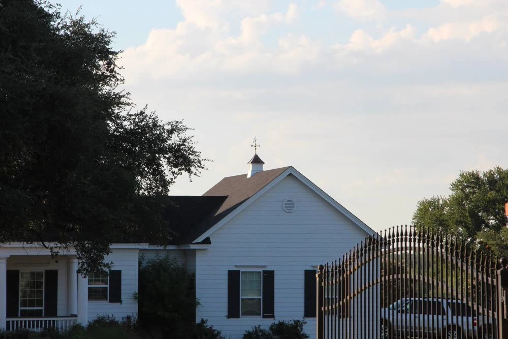 A white house with a cupola and tall trees, before roofing work