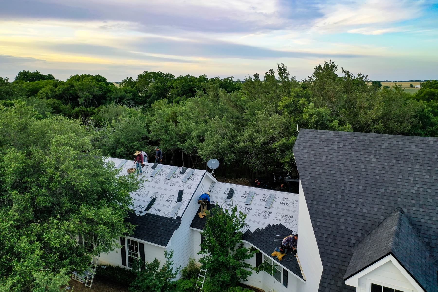 A Hardtop crew laying felt on a white house in Central Texas, with a finished shingle roof beside it
