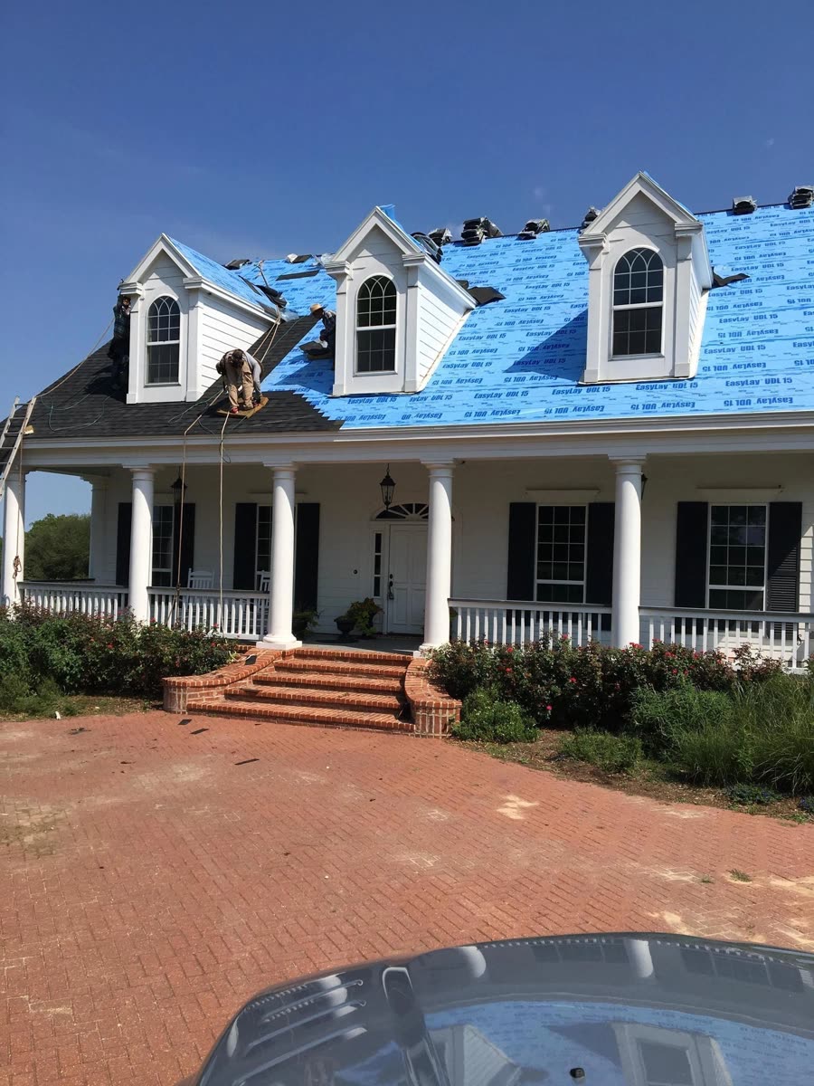 A brick-red drive in front of a two-story house with blue felt on the roof and dormer windows